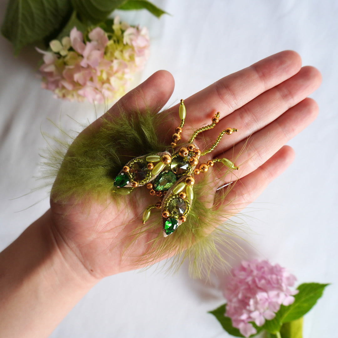 Hand holding a decorative  fly TGbrooch with green stones and gold accents
